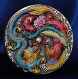 A round silver-toned metal medallion featuring a detailed relief design of a blue dragon and a red phoenix surrounding a pink peony, set against a dark blue satin background.