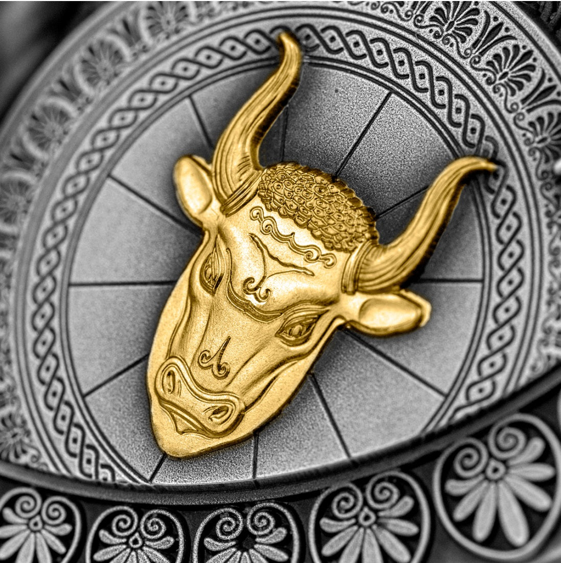 Extreme close-up photo of the high-relief shield detail on the antique silver coin. The shield features a highly detailed, fierce bull's head sculpted  and gilded in gold creating a stark contrast against the dark, oxidized background of the antique silver coin's surface.