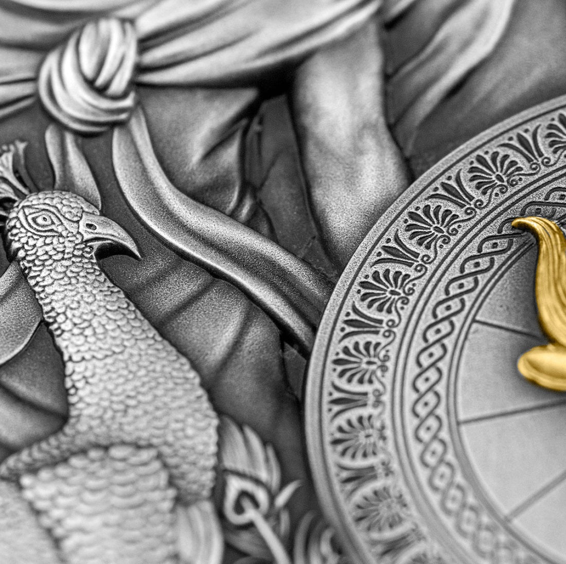 A close-up photo of the antique silver coin focusing on Juno's attributes. It shows the detailed, cascading feathers of a peacock layered next to the high-relief shield, which shows the horn of a  features a powerfully sculpted bull's head with gold plat.ng.