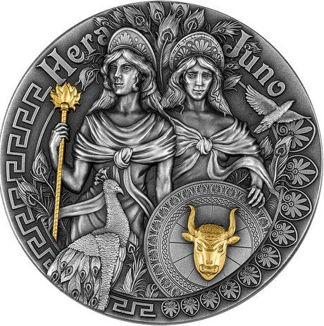 A circular silver coin featuring relief images of Hera and Juno with a gilded finish of a flower topped staff and a bull's head on a shield. Greek classical patterns adrn the edge of the face of the coin.