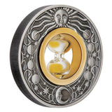 An angled view of a round silver-toned medallion or coin featuring a sun face at the top, moon phases around the border, and a central gold-toned oval with an hourglass.  A reeded rim is evident.