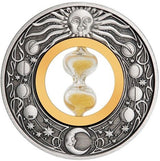 A circular silver coin with an antiqued finish, featuring a central hourglass with golden sands and astronomical symbols around it. A round silver-toned medallion or coin featuring a sun face at the top, moon phases around the border, and a central gold-toned oval with an hourglass. The piece is photographed on a dark blue satin-like background.