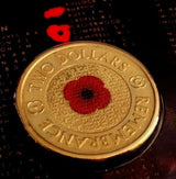 Coin close-up: Australian $2 Remembrance coin with red poppy centre, fine background lettering and C mintmark, photographed against a dark poppy background.